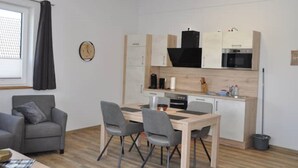 Un espace de vie compact comprenant un coin cuisine, un coin repas avec une table et des chaises, un canapé gris et une horloge au mur.