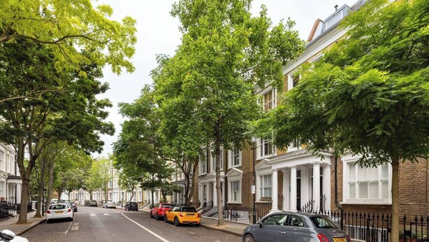 Une rue bordée d’arbres, avec des voitures garées et des maisons traditionnelles en briques.