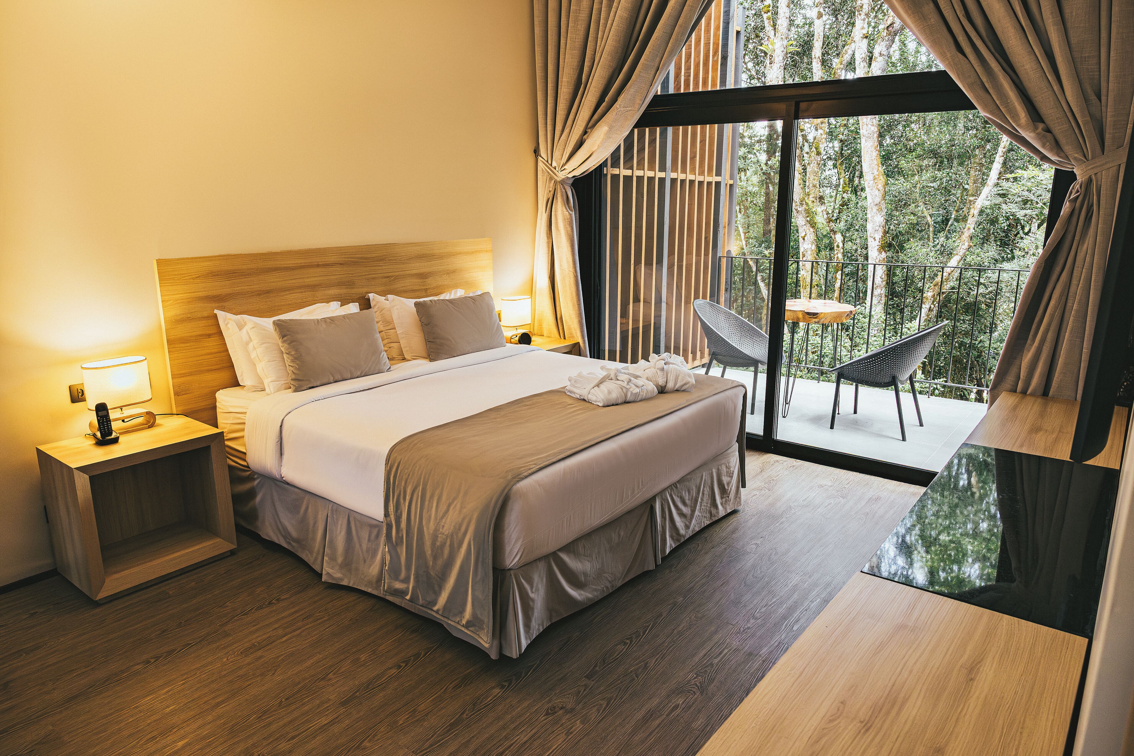 Habitación de hotel moderna con una cama grande, mesitas de noche con lámparas, un balcón con una silla y vistas a árboles por la ventana.