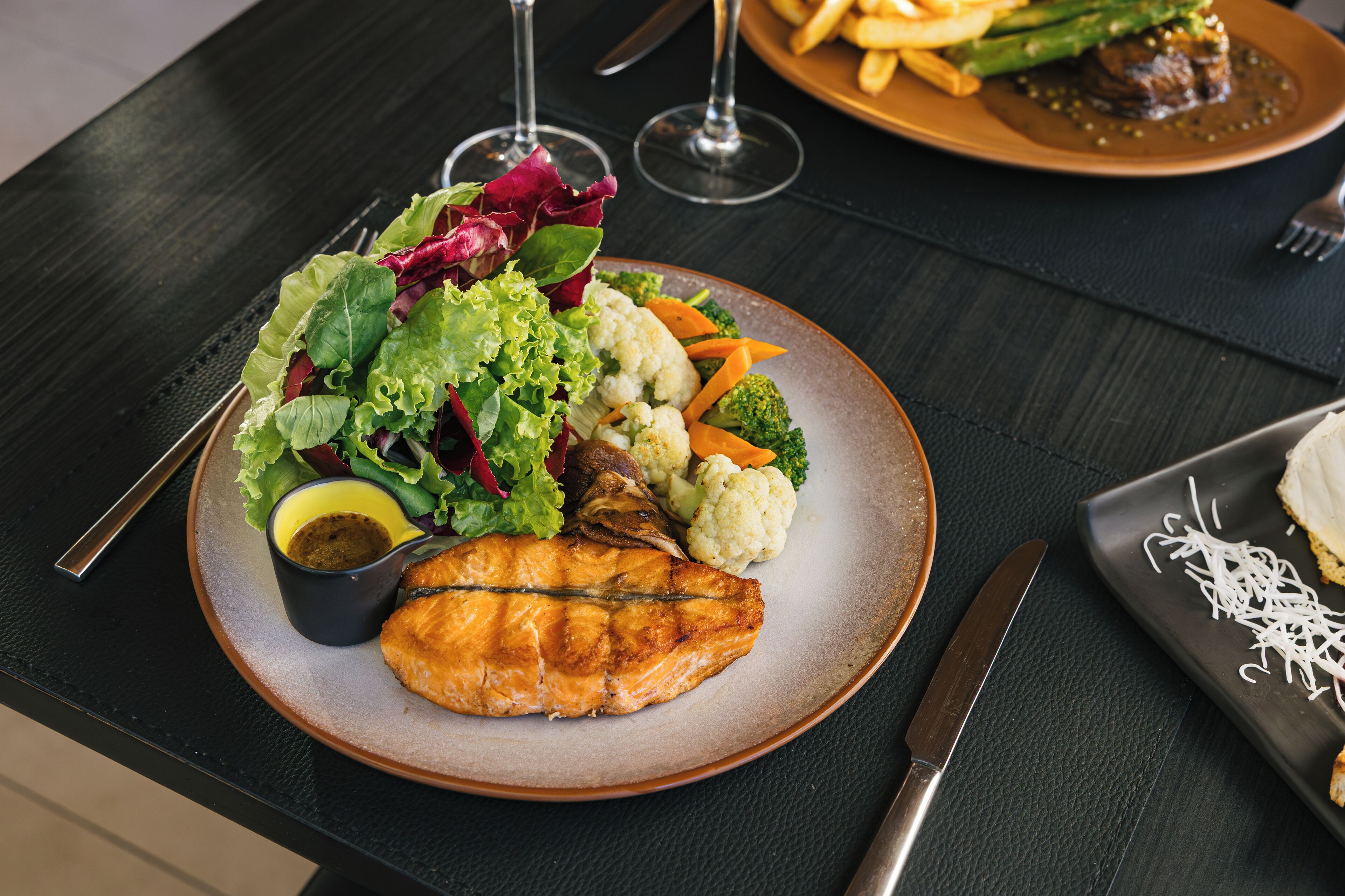 A plate of food with a piece of grilled fish, vegetables, and a small bowl of sauce.