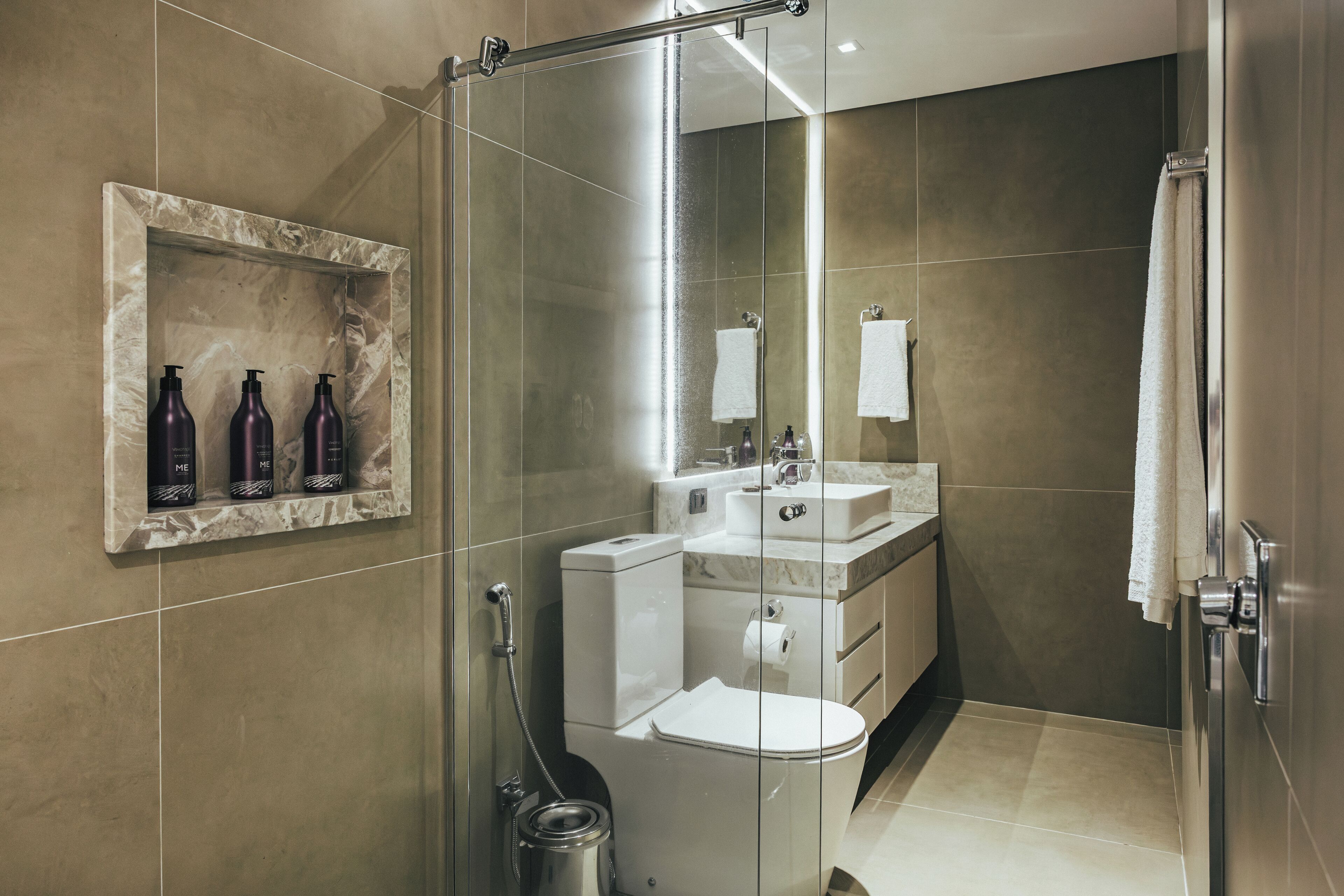 Un baño moderno con ducha de vidrio, estante de mármol con artículos de aseo, tocador con espejo y retrete.