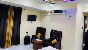 A hotel room with a bed, two armchairs, a small table, a TV, an air conditioning unit, and a ceiling fan. - Hotel Almarkaz (Islamabad)