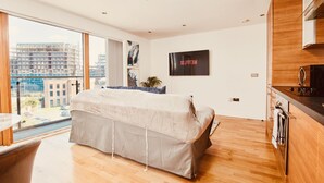 A modern bedroom with a large bed, a view of the city, and a kitchenette.