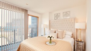 A hotel room with a large bed, two bedside tables with lamps, a vase with flowers, and a view of the city through floor-to-ceiling windows.