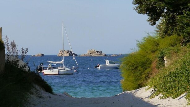 Escena costera con barcos anclados cerca de una playa de arena, rodeada de vegetación y afloramientos rocosos.
