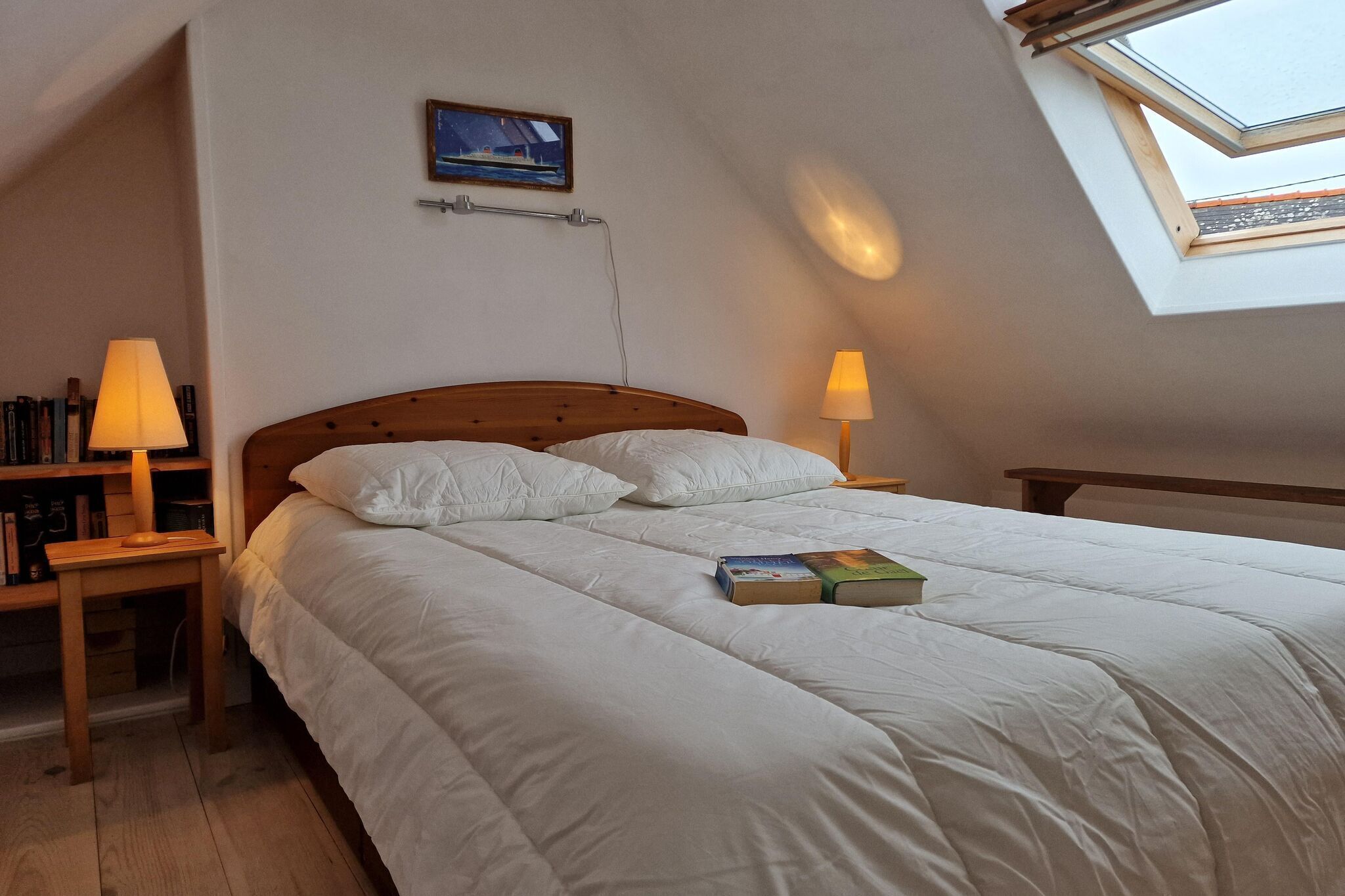 A bedroom with a wooden bed, two bedside lamps, a bookshelf, and a skylight.