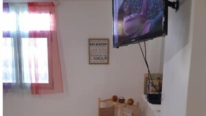 Quarto com televisão fixada na parede, prateleira de madeira com diversos objetos, cesta de vime e janela com cortinas.