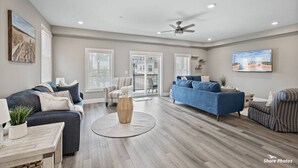 Ein geräumiges Wohnzimmer mit einem großen blauen Sofa, einem gestreiften Sessel und einem Holztisch. Zwei Fenster mit Jalousien, ein Deckenventilator und ein Gemälde an der Wand sind ebenfalls vorhanden.