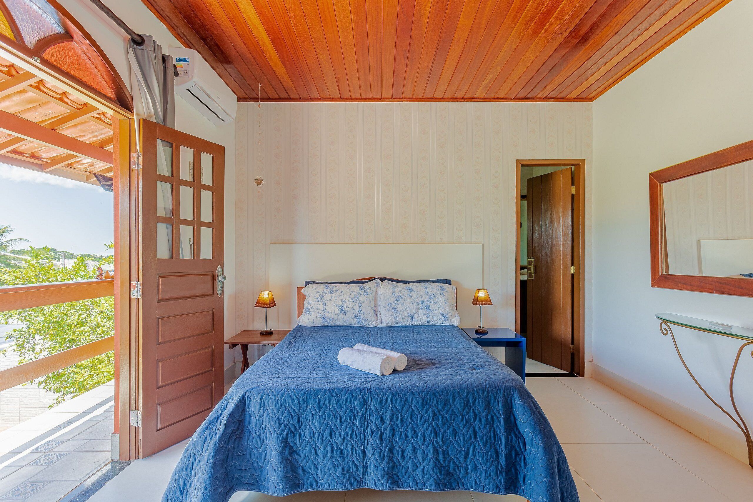 Quarto com teto de madeira, cama com colcha azul, duas luminárias de cabeceira e vista para o exterior.