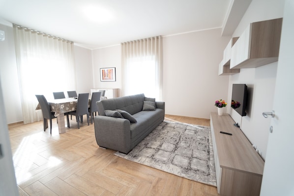 Un salon moderne avec un canapé gris, une table à manger avec des chaises, un téléviseur posé sur un meuble en bois et un tapis à motifs.
