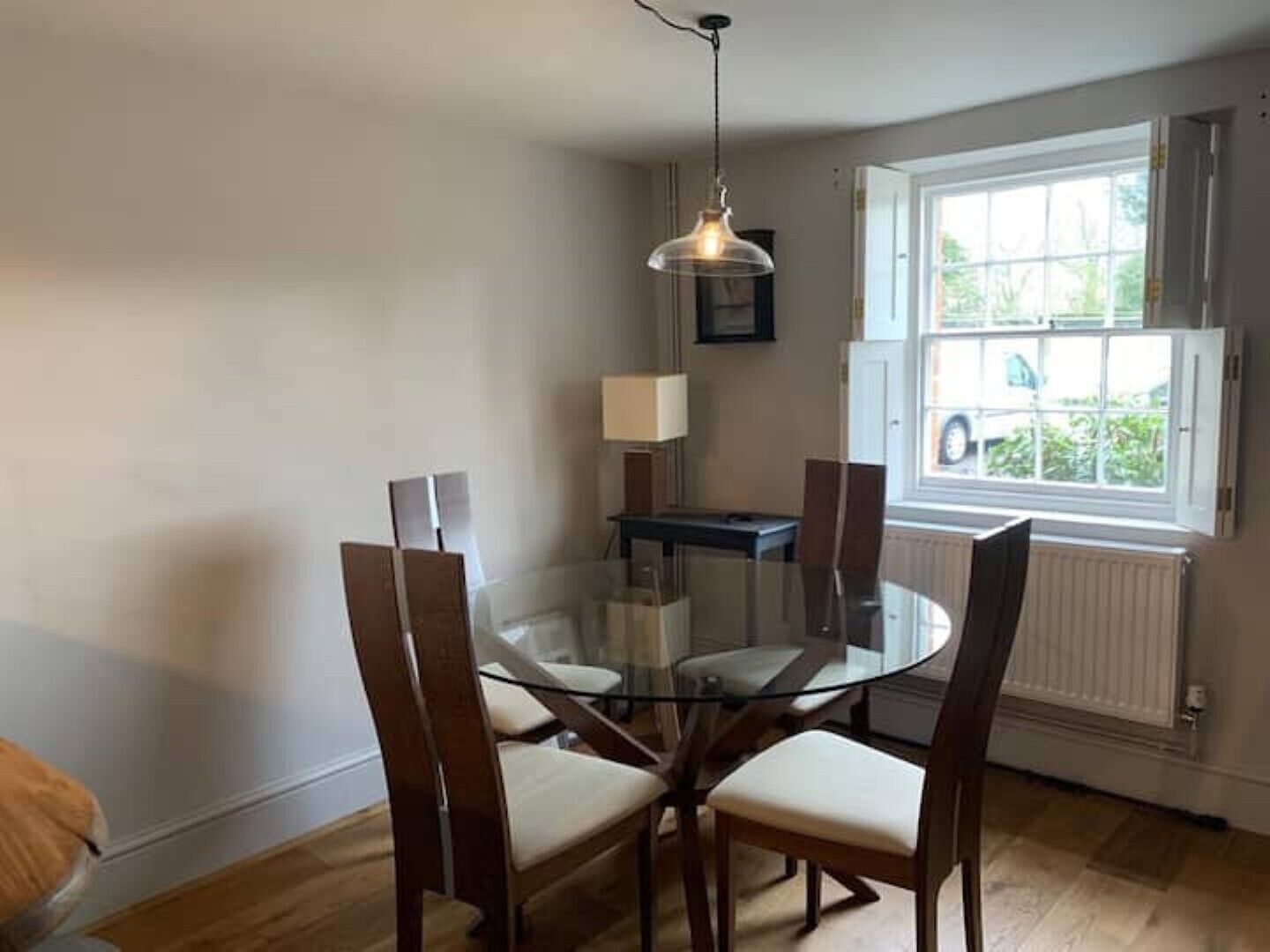 A dining area with a glass table and wooden chairs, a pendant light, a window with a view of greenery, and a radiator.