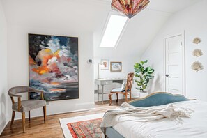 A bedroom with a large abstract painting, a desk, a chair, a bed, and a skylight.