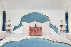 A bed with a blue headboard, white bedding, and a red patterned pillow. There are two bedside lamps with gold frames.