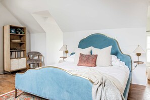 A bedroom with a blue upholstered bed, a wooden bookshelf, a chair, and two bedside lamps.