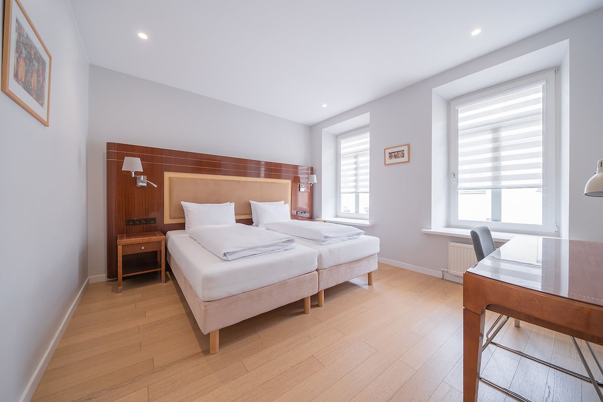 A bedroom with a wooden headboard, two beds, bedside tables, a desk, and a window with blinds.