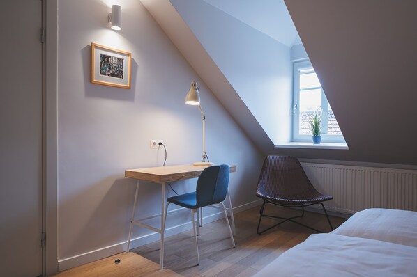 A bedroom with a sloped ceiling, a desk with a chair, a bedside table, and a window with a view of the outside.