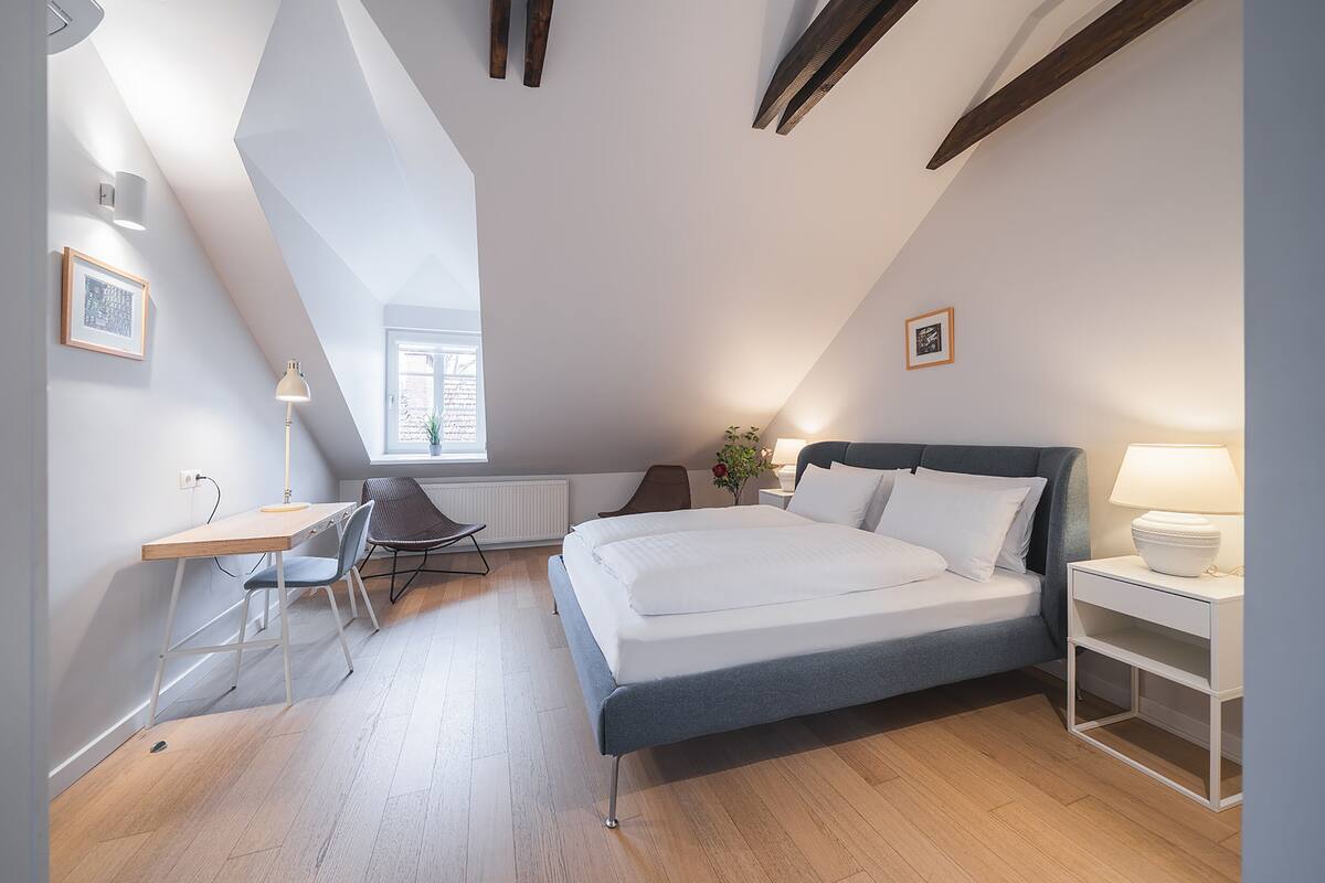 A modern bedroom with a sloped ceiling, a bed, a desk, a chair, and a nightstand.