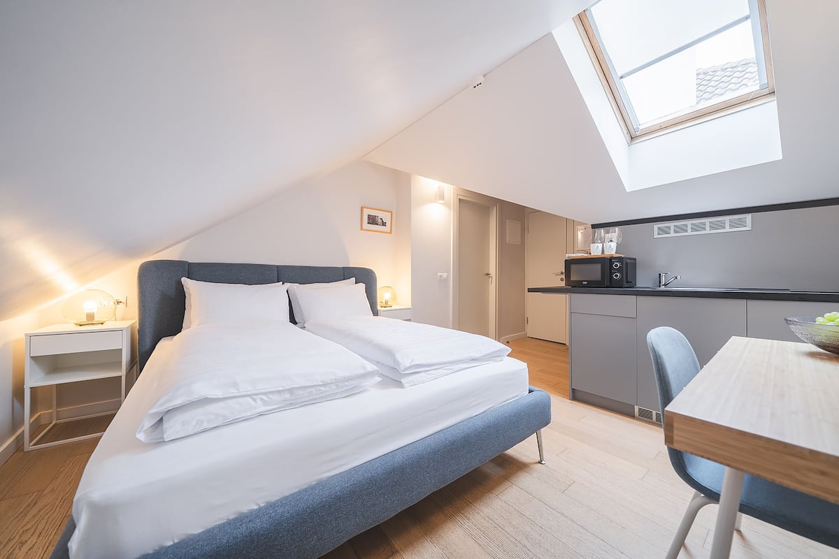 A modern bedroom with a skylight, a bed, a bedside table, a desk, and a kitchenette.