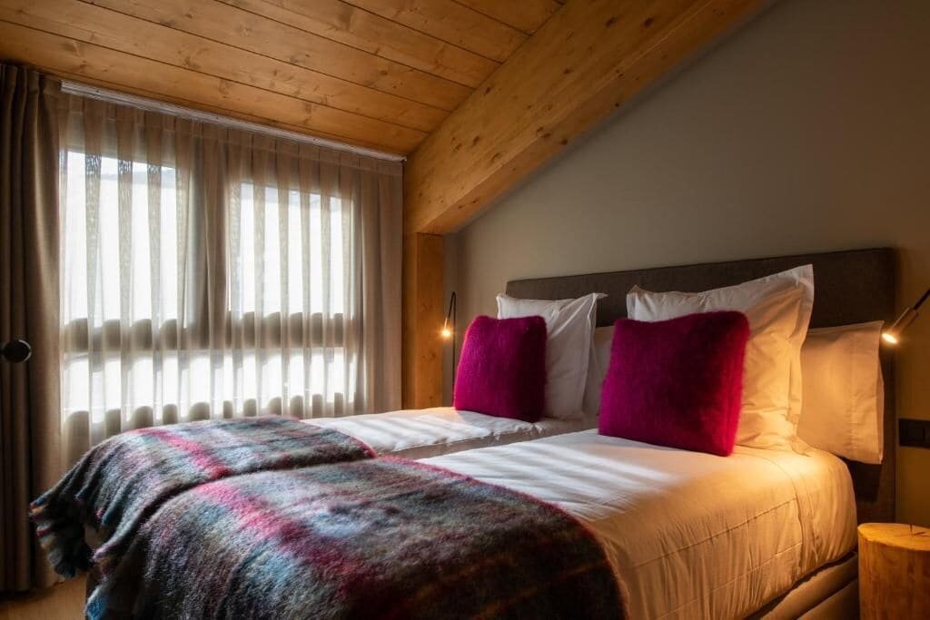 A cozy bedroom with a wooden ceiling, a large bed with white linens and two red pillows, and a window with sheer curtains.