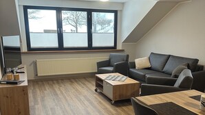 Un salon moderne avec un canapé, une table basse et un meuble pour la télévision. Il y a une grande fenêtre avec des stores, un radiateur et un luminaire au plafond.