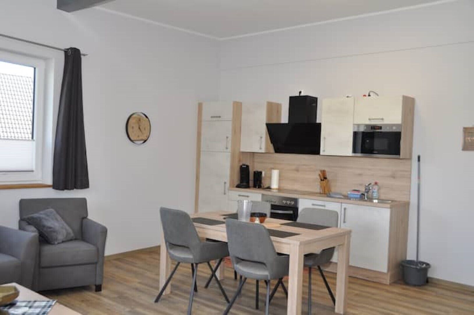 A compact living space with a kitchenette, a dining area with a table and chairs, a gray sofa, and a clock on the wall.