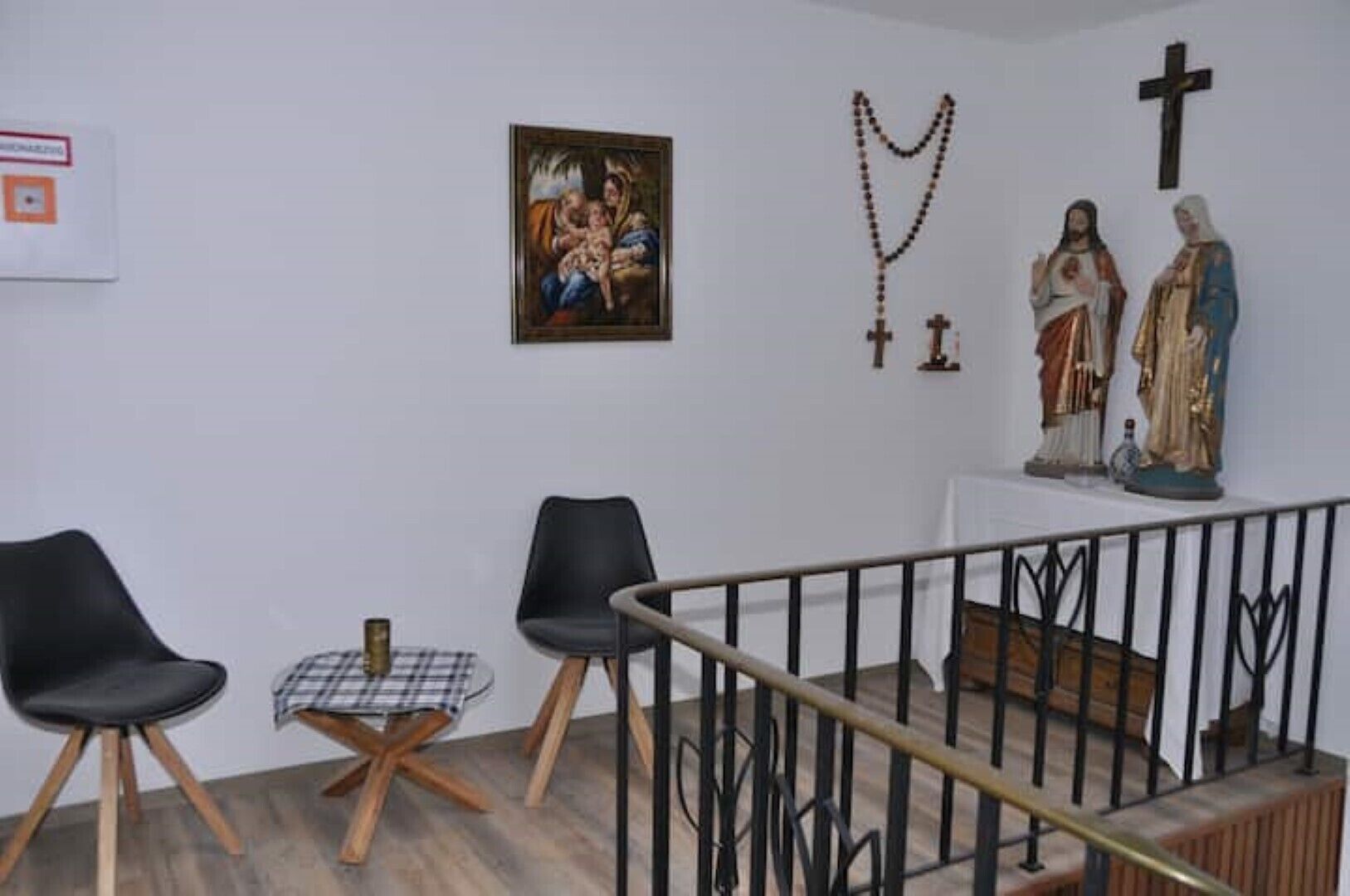 A room with a white wall, a painting, a rosary, and two statues. There are two chairs and a small table.