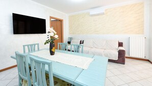 A dining area with a table, chairs, a vase with flowers, a flat-screen TV, a sofa, and a radiator.