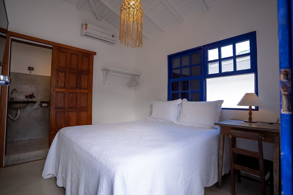 A bedroom with a bed, a wooden door, a window with blue trim, a bedside table with a lamp, and an air conditioning unit on the wall.