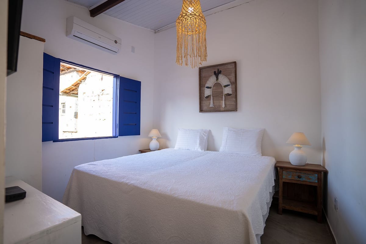 A bedroom with a bed, two bedside lamps, a wall-mounted air conditioner, a window with blue shutters, and a hanging macrame light fixture.