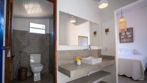 Een badkamer met toilet, wastafel en douche. In de aangrenzende kamer staat een bed met wit beddengoed.