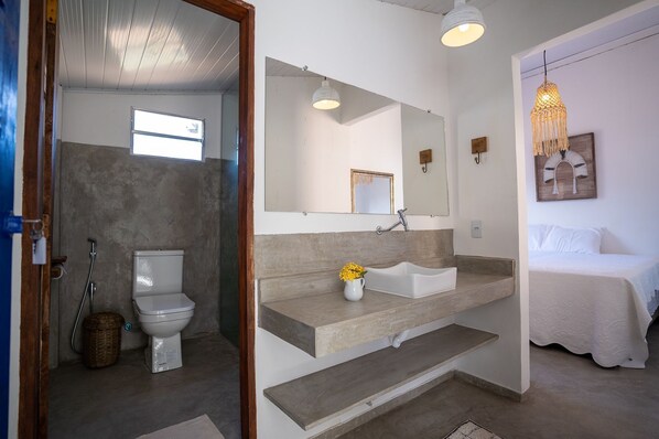 A bathroom with a toilet, sink, and shower. There is a bed with white bedding in the adjacent room. - Arandu Casa Hotel Quadrado (Porto Seguro)