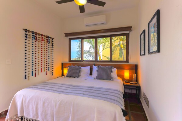A bedroom with a large bed, a ceiling fan, a window with a view of palm trees, and a wall-mounted air conditioner.