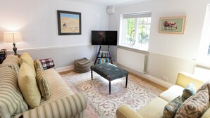 A living room with a sofa, ottoman, and a flat-screen TV. There is a bookshelf and a window with a view of greenery.