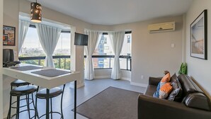 Ein modernes Wohnzimmer mit Küchenbereich, großen Fenstern mit Vorhängen, einem braunen Ledersofa und einem fest montierten Flachbildfernseher.