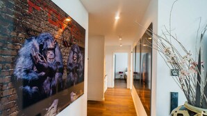 A hallway with a brick wall mural, wooden flooring, and a large window with a view of a room.