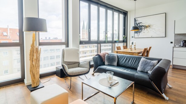 A modern living room with a black leather sofa, a wooden coffee table, and a floor lamp. Large windows offer a view of the cityscape.