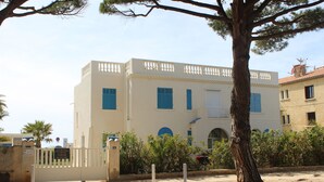 A two-story building with a white facade and blue shutters, surrounded by a gated entrance and a large tree.