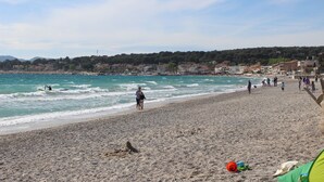 A beach with people walking along the shore, a clear sky, and buildings in the background.