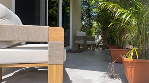 Un espace extérieur moderne aménagé avec un canapé et des fauteuils, entouré de plantes en pot et doté d’une porte vitrée donnant sur un jardin.