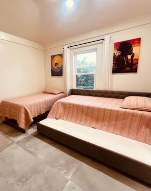 Une chambre avec deux lits simples et un lit plus grand, une fenêtre avec des rideaux et des œuvres d’art accrochées aux murs.