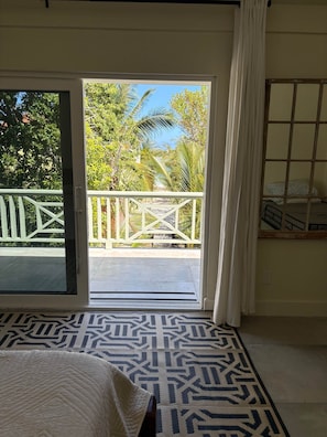 A room with a bed, a patterned rug, a view of a balcony with a railing, and a window with a curtain.