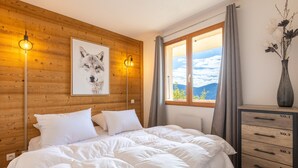A bedroom with a wooden headboard, a large bed with white bedding, a framed picture on the wall, and a window with a view of the sky and mountains.