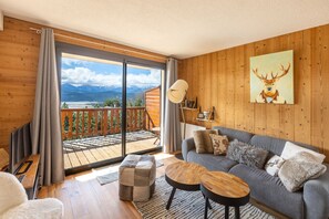 A cozy living room with a grey sofa, wooden coffee table, and a view of mountains through a sliding glass door. - Le Reflet du Lac - Station de ski accessible à pied (Les Angles)
