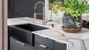 Una cucina moderna con un piano di lavoro in marmo, mobili neri, un rubinetto dorato e una lampada a sospensione.
