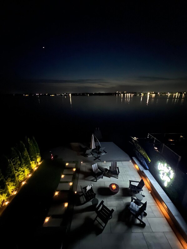 An outdoor patio with lounge chairs and a fire pit, overlooking a calm body of water at night.