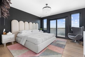 A modern bedroom with a large bed, a nightstand, a chair, and a view of the outdoors through large windows.