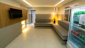 A modern hotel lobby with a flat-screen TV mounted on a wooden wall, a seating area with a vase of yellow flowers, and a refrigerator stocked with beverages. - Hotel Pousada do Farol (Aracaju)