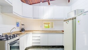 A kitchen with white cabinets, a stove, a refrigerator, and a window with a view of greenery. - GB69 Excelente Casa a 400m da Praia (Camaçari)