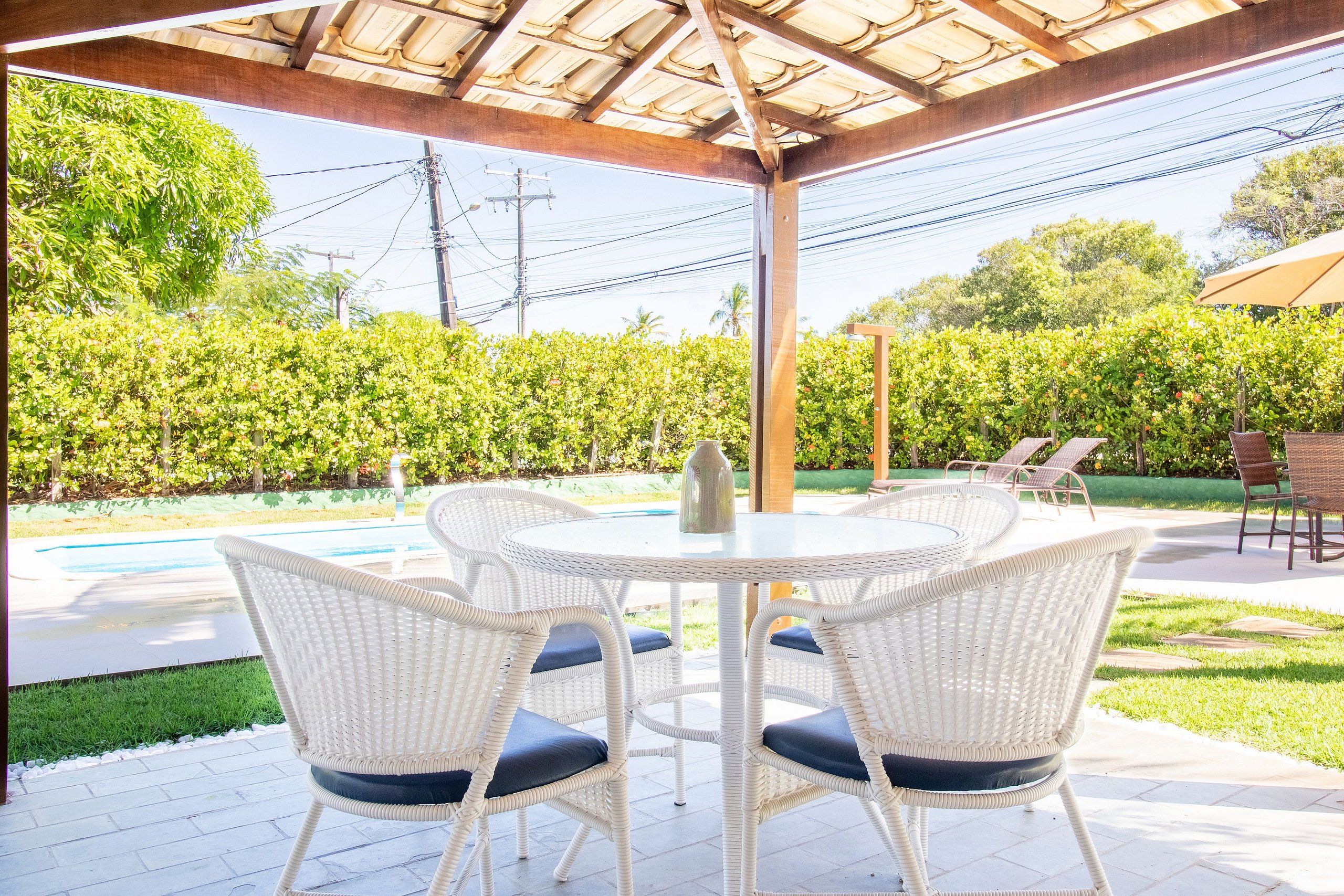 A covered patio with a round table and white wicker chairs, overlooking a swimming pool and lush greenery.