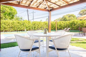 A covered patio with a round table and white wicker chairs, overlooking a swimming pool and lush greenery. - GB69 Excelente Casa a 400m da Praia (Camaçari)